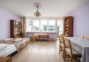 A room with two beds, a dining table, chairs, a bookshelf, and a window with a view of buildings. - Złota Spacious Apartment (Warszawa)