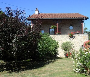 Edifici de pedra amb teulada a dues aigües, envoltat d'un jardí ben cuidat amb diverses plantes i flors.