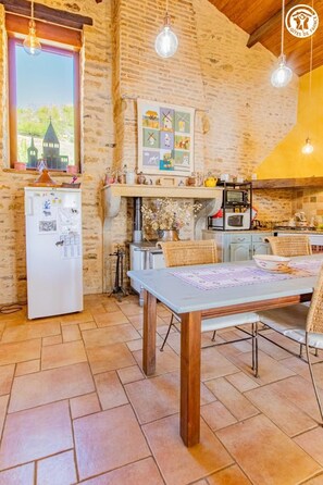 Cuina rústica amb paret de maó, nevera, taula de dinar amb caires i lluminàries penjants.