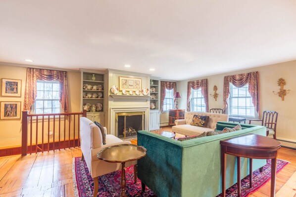 Een ruime woonkamer met een open haard, een groene bank, een witte fauteuil en een kleine ronde tafel.