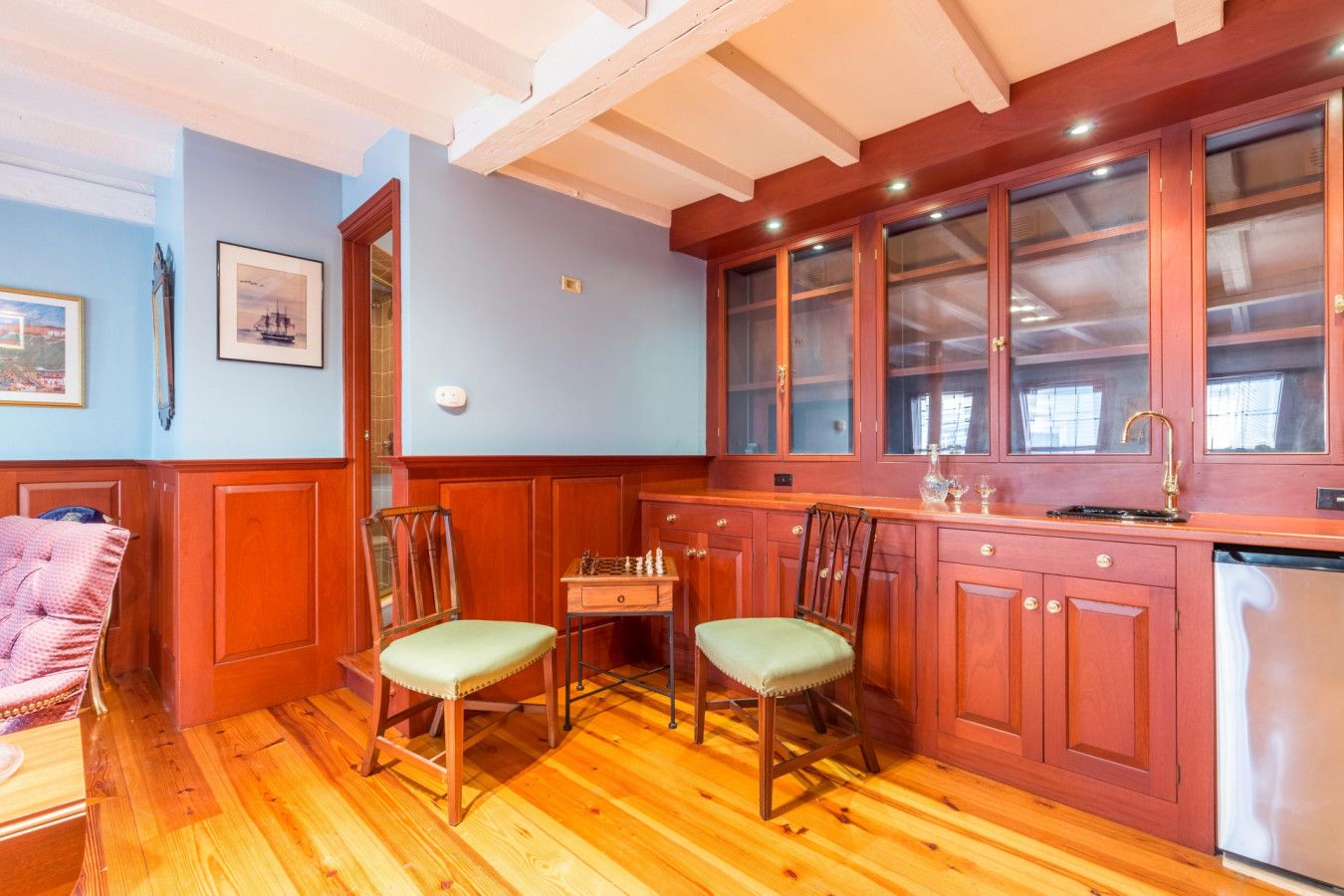 A room with wooden flooring, a dining table with chairs, a kitchen area with cabinets and a sink, and a framed picture on the wall.