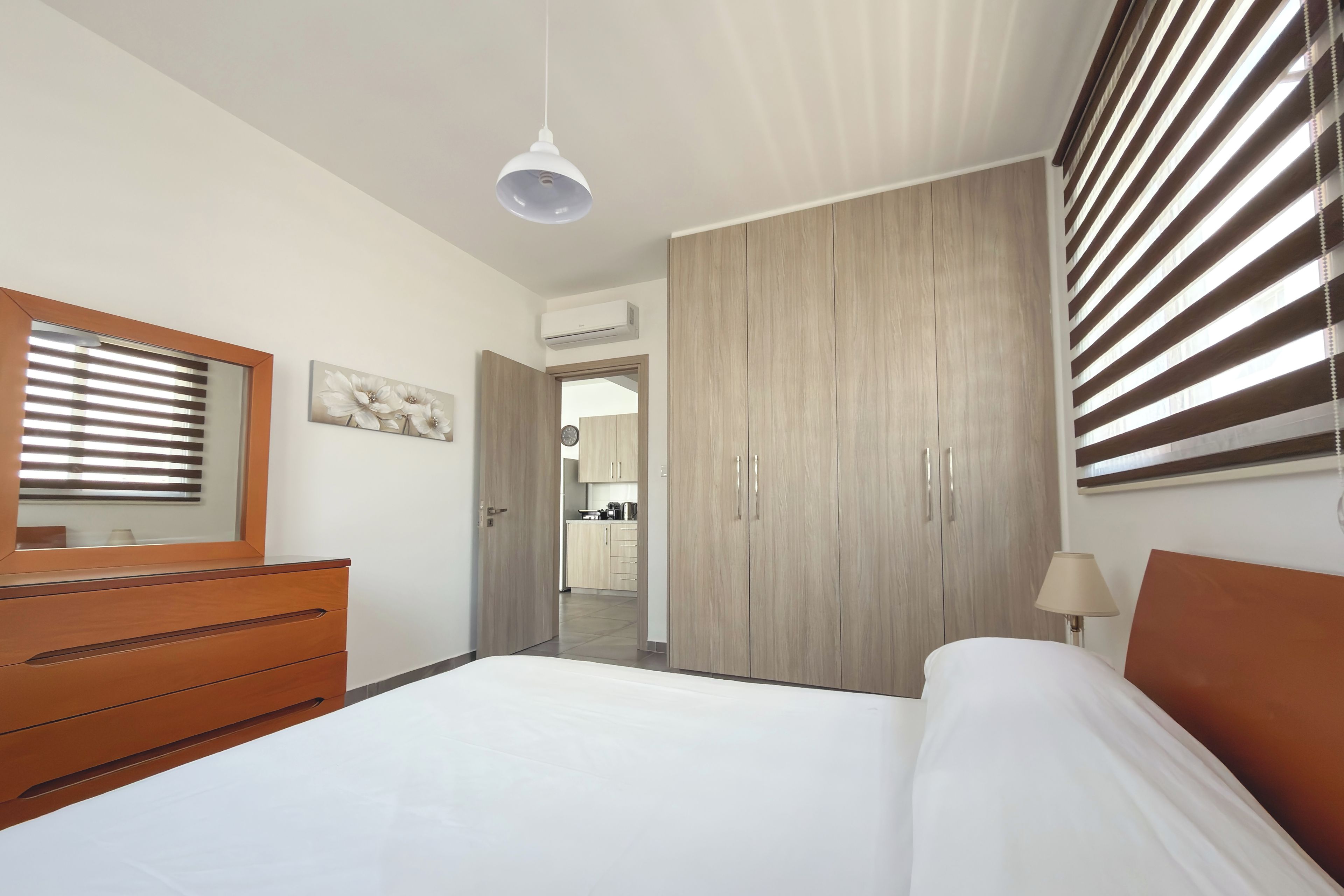 Un dormitorio con cama, cómoda, espejo, armario, lámpara de techo y ventana con persianas.