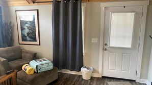 Ein Zimmer mit einer Couch, einem gerahmten Bild, einer Tür mit Fenster und einem Mülleimer.