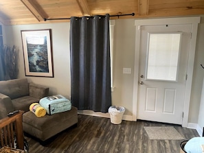 Una habitación con un sofá, un cuadro enmarcado, una puerta con ventana y un cubo de basura.