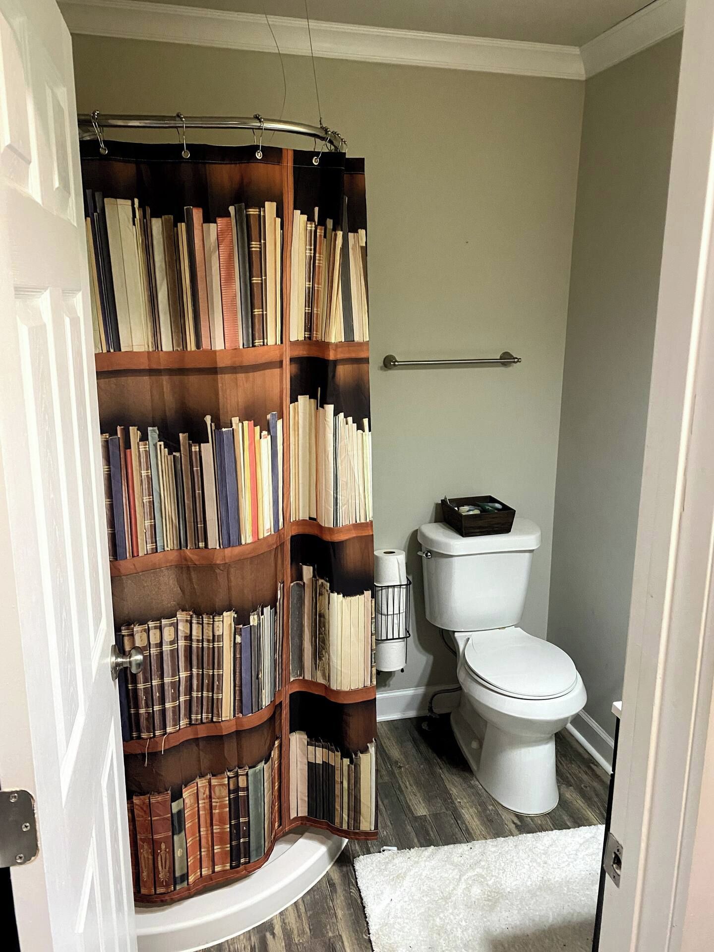 A bathroom with a wooden bookshelf filled with books, a white toilet, and a shower curtain.