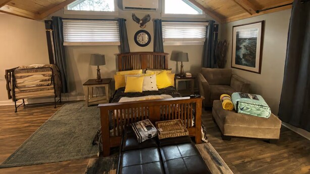 Ein gemütliches Schlafzimmer im Loft-Stil mit schrägem Dach, einem Bett mit gelber Bettwäsche, einer hölzernen Bank, einem Sofa und einem gerahmten Bild an der Wand.