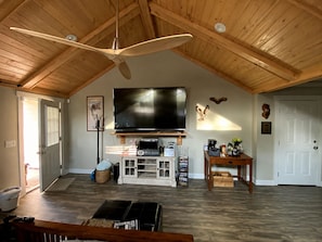 Una sala de estar amplia con techo alto, vigas de madera, un televisor de pantalla plana grande, una chimenea y una mesa de centro.