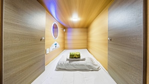 A small, enclosed room with a bed, a circular mirror, and a control panel on the wall. - Sleep Different-Stay Inspired-Where inovation meets coastal living-Capsule Hotel (Benowa)