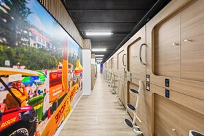 A row of wooden lockers with numbered doors, a tiled floor, and a corridor with a beach-themed mural on the left. - Sleep Different-Stay Inspired-Where inovation meets coastal living-Capsule Hotel (Benowa)