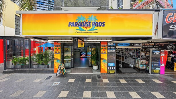 A storefront with a sign reading 'Paradise Pods' and a sign for 'Oz Blasters' across the street. - Sleep Different-Stay Inspired-Where inovation meets coastal living-Capsule Hotel (Benowa)