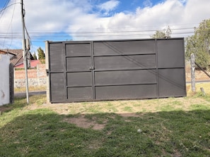 Une grande porte en métal, composée de plusieurs panneaux, est installée dans une zone herbeuse, avec un mur de briques et un bâtiment à toit rouge en arrière-plan.