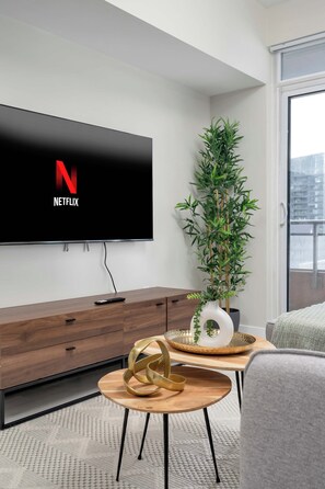 Ett modernt vardagsrum med en platt-TV som är monterad på väggen och visar Netflix-logotypen, ett träskåp med en fjärrkontroll, en hög grön växt och ett runt träbord som används som soffbord, med en dekorativ detalj på.