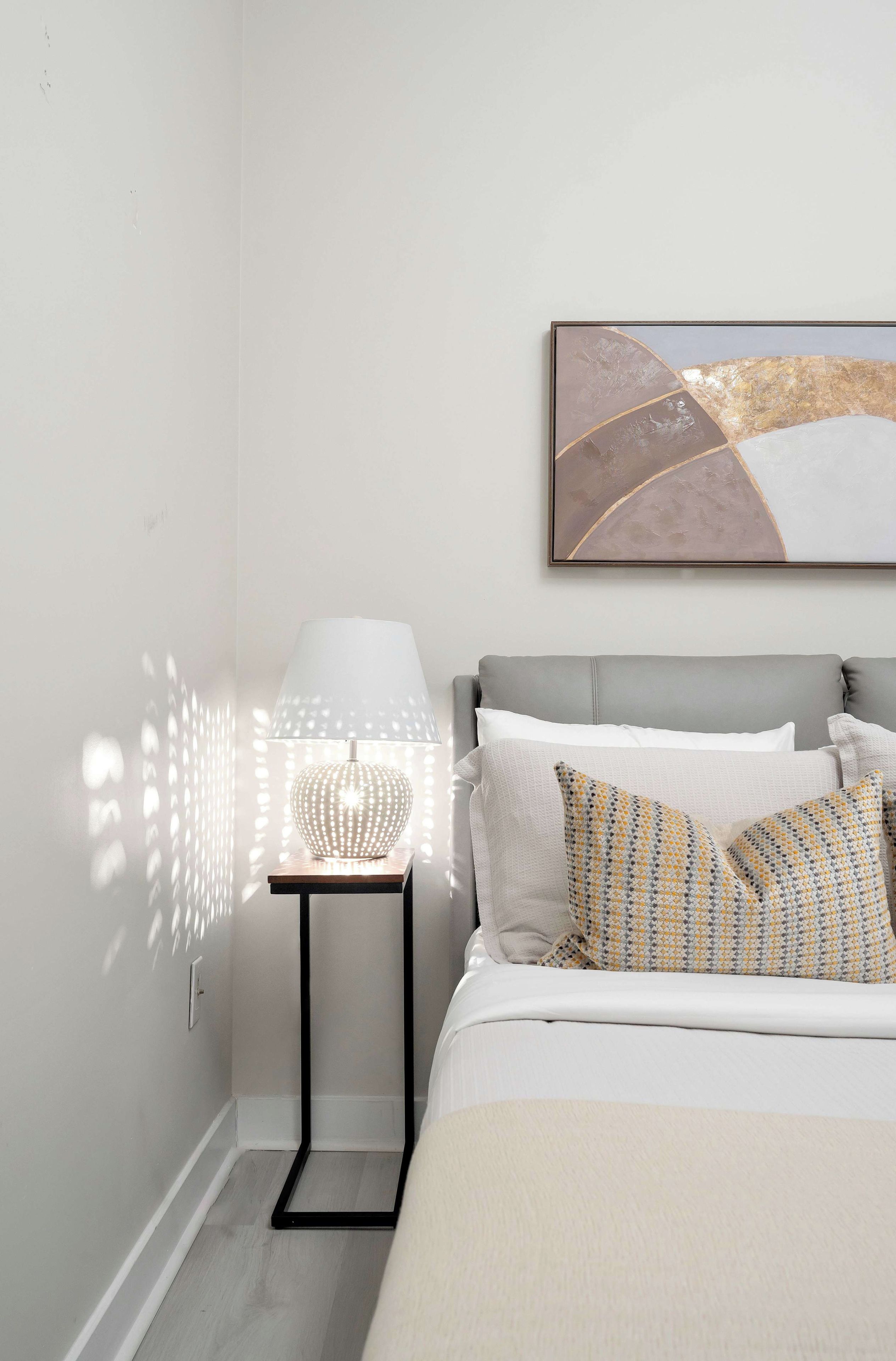 A bedroom with a bed, a bedside table with a lamp, and a wall-mounted abstract painting.