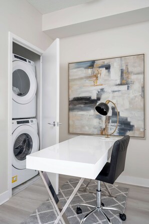 A room with a white table, a black office chair, a floor lamp, a painting, and a washing machine. - Lakeview CityPlace 2BR | Free Parking & Balcony | Parking Available (Toronto)