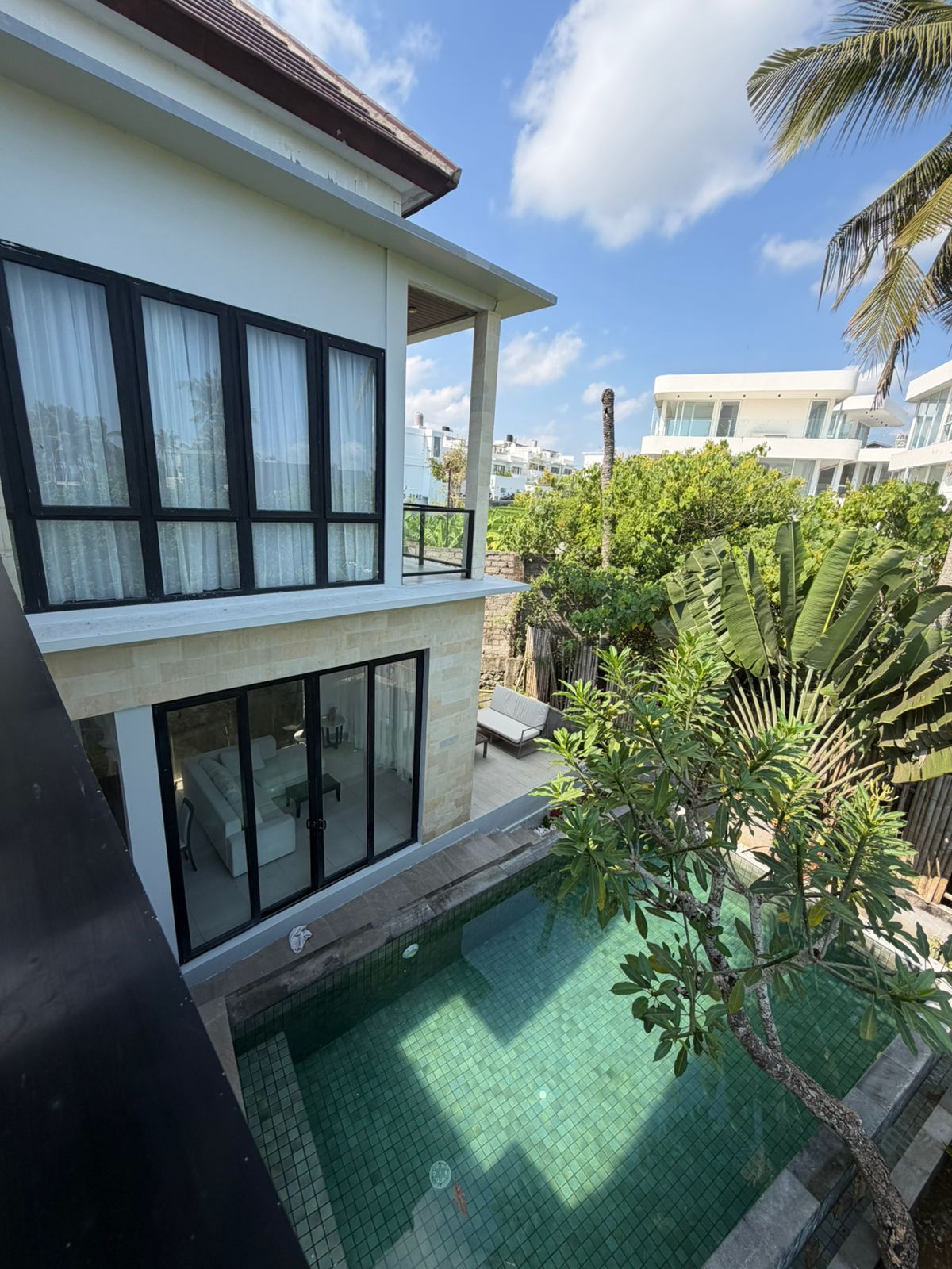 Un edificio moderno de dos pisos con piscina, mobiliario exterior y abundante vegetación.