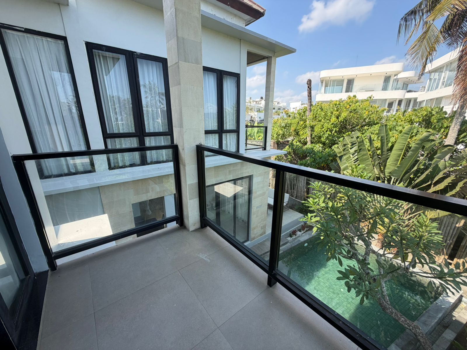 A balcony with glass railings, a tiled floor, and a view of a swimming pool and greenery.