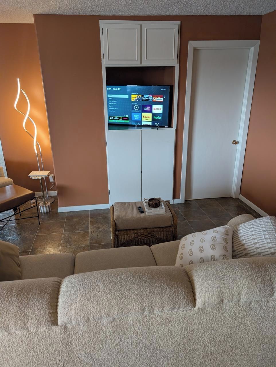 Una sala de estar con un televisor de pantalla plana montado en un mueble blanco, una lámpara de pie, un sofá marrón y una mesita con un control remoto.