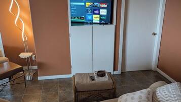 Una sala de estar con un televisor de pantalla plana montado en un mueble blanco, una lámpara de pie, un sofá marrón y una mesita con un control remoto.