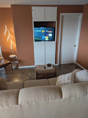 A living room with a flat-screen TV mounted on a white cabinet, a floor lamp, a brown sofa, and a small table with a remote control.