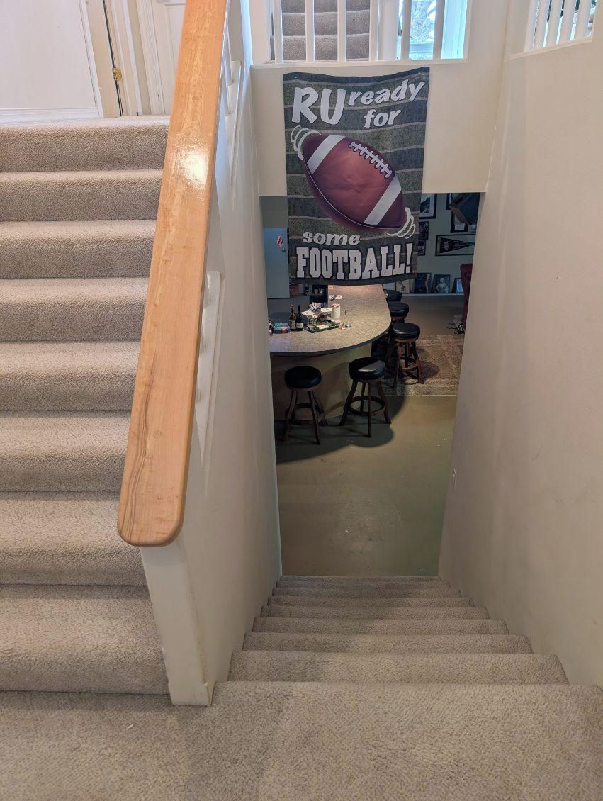 A staircase with a wooden handrail leading to a landing with a kitchen area, a bar stool, and a poster that reads "RU ready for some FOOTBALL!"