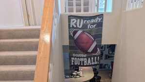 A staircase with a wooden handrail leading to a landing with a kitchen area, a bar stool, and a poster that reads "RU ready for some FOOTBALL!" - Bed and Breakfast • King Bed + Game Room + Fire Pit (Kiron)