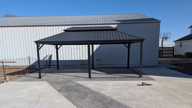A covered patio area with a metal roof and concrete flooring, adjacent to a white building with a corrugated metal exterior. - Bed and Breakfast • King Bed + Game Room + Fire Pit (Kiron)