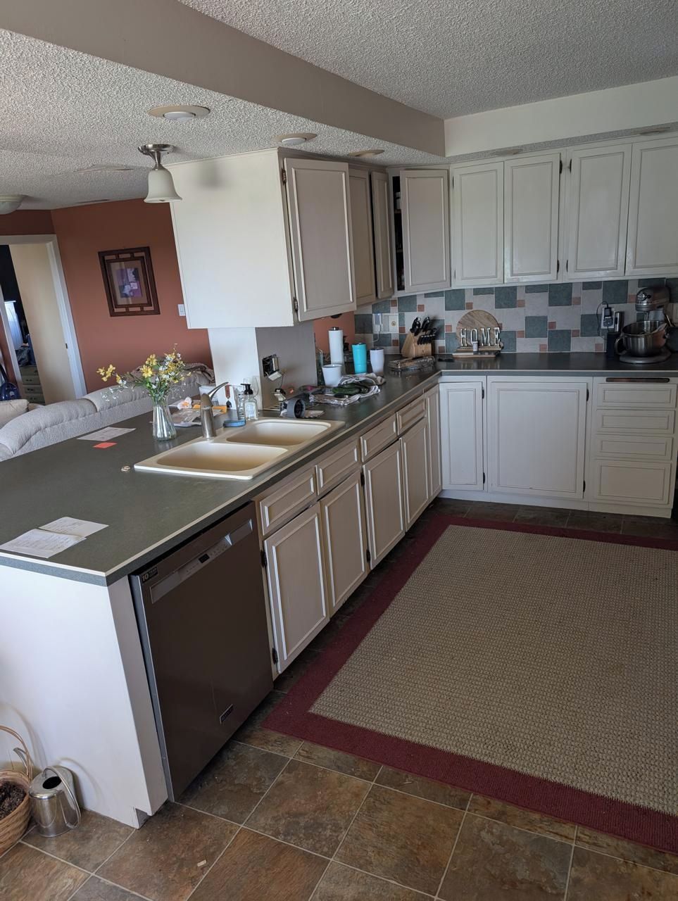 A kitchen with white cabinets, a stainless steel dishwasher, a tiled backsplash, and a red rug.