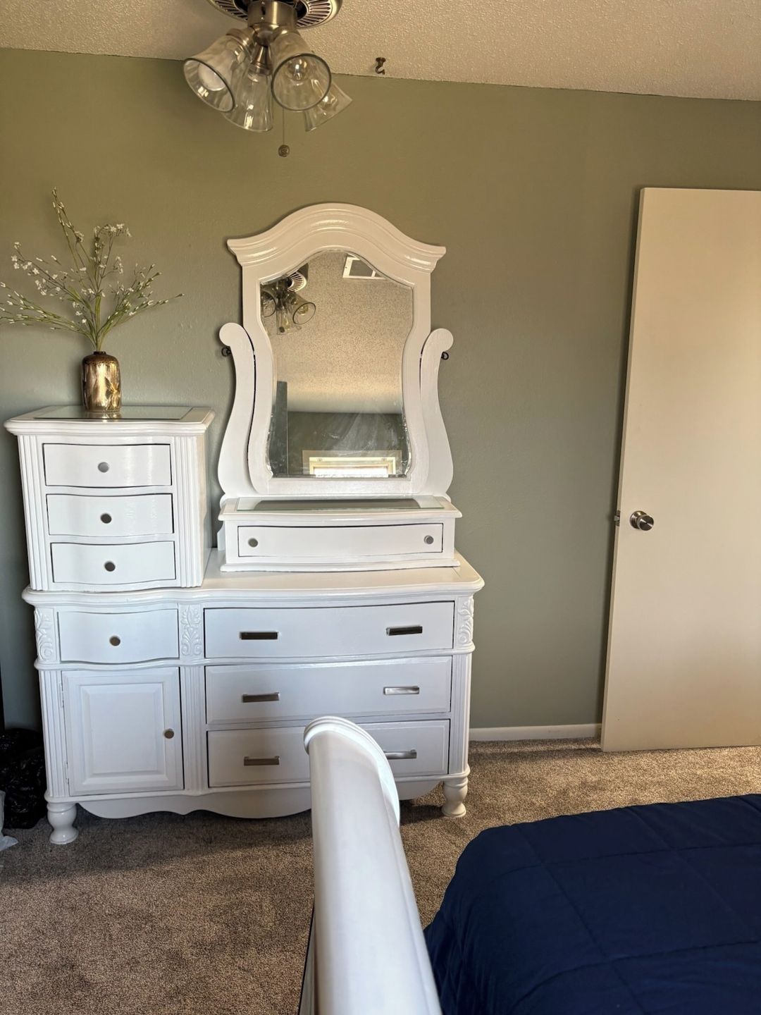 A bedroom with a white dresser, a mirror, a vase with flowers, and a bed with a blue cover.