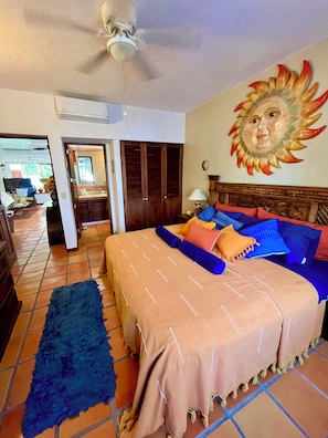A bedroom with a bed, a ceiling fan, a wall mural, and a view into another room.
