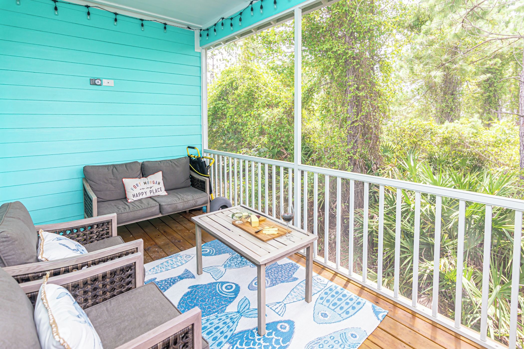 A covered porch with a turquoise wall, a gray sofa with a pillow, a wooden table with a fish-patterned rug, and a railing overlooking a dense green forest.