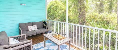 A covered porch with a turquoise wall, a gray sofa with a pillow, a wooden table with a fish-patterned rug, and a railing overlooking a dense green forest.