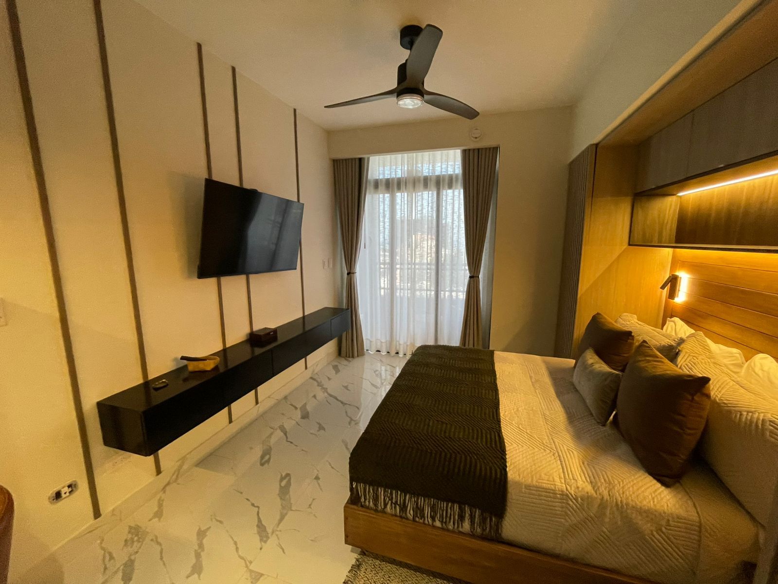 A modern bedroom with a bed, a TV mounted on the wall, a ceiling fan, and a bedside table.