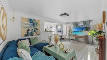 Ein Wohnzimmer mit einem blauen Sofa, einem grünen Tisch und einem Fernseher, der eine Strandlandschaft zeigt.