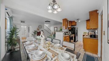 Comedor con una larga mesa dispuesta para una comida, una cocina al fondo y una escalera que sube a otro nivel.