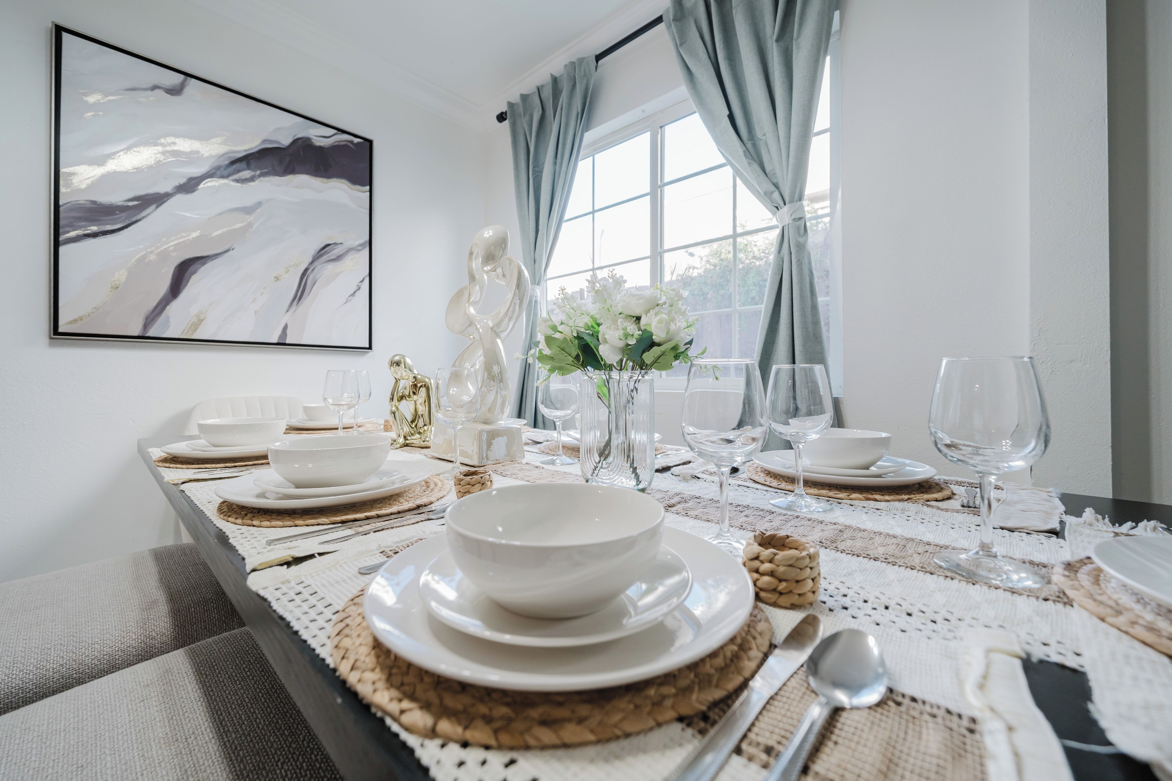 A dining table set with white plates, bowls, and cutlery, with a vase of white flowers and a decorative sculpture.
