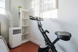 Habitació amb una bicicleta d'exercici vertical, un aire fresc blanc, una estanteria blanca amb una planta i una obra d'art emmarcada a la paret.