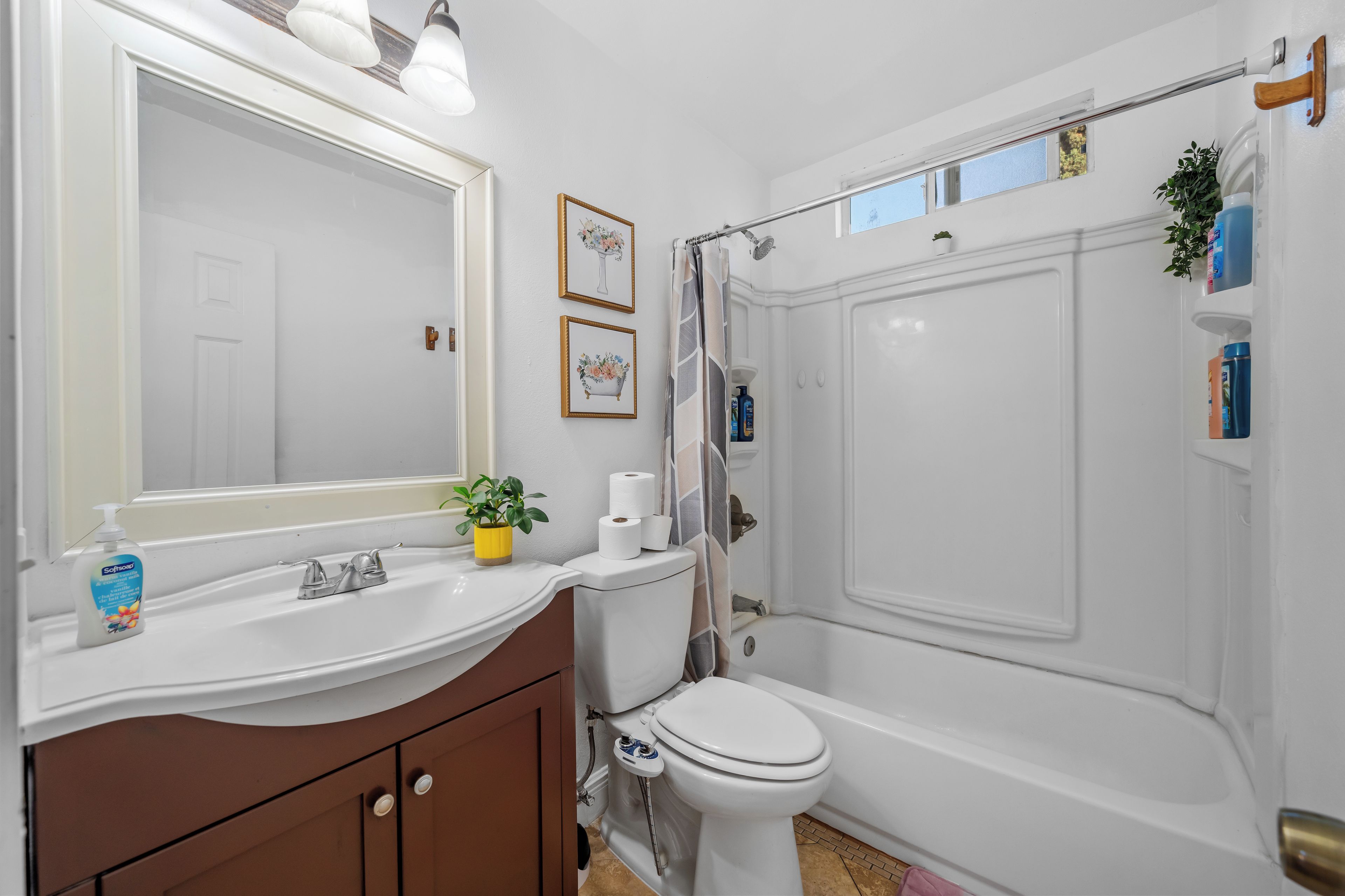 Ein Badezimmer mit Waschbecken, Toilette und Badewanne. An den Wänden hängen Bilder in Rahmen, auf dem Waschbecken steht ein Topf mit einer Pflanze und auf der Ablage liegen Pflegeprodukte.