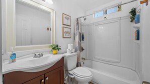 A bathroom with a white sink, toilet, and bathtub. There are framed pictures on the wall, a potted plant on the sink, and toiletries on the countertop. - Garage, Gated Yard, near SD Gaslamp, DT, 4BR/2Bth (National City)