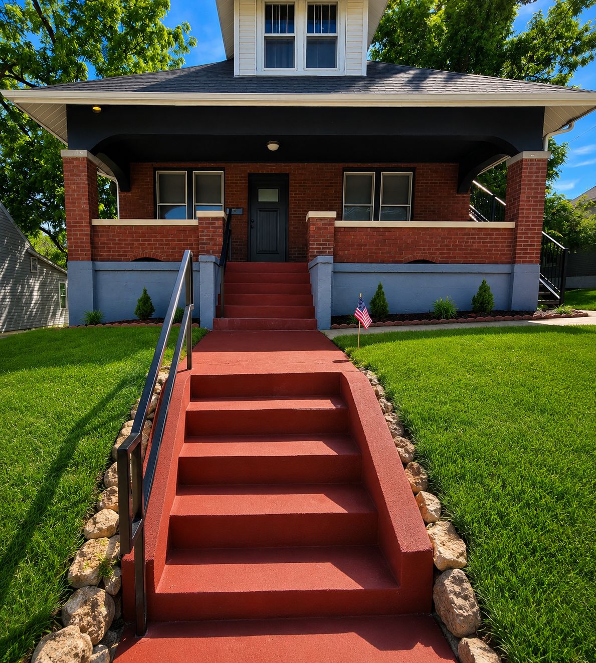Une maison à deux étages avec un porche rouge à l’avant, un escalier menant à la porte d’entrée et un petit drapeau américain planté dans la pelouse.