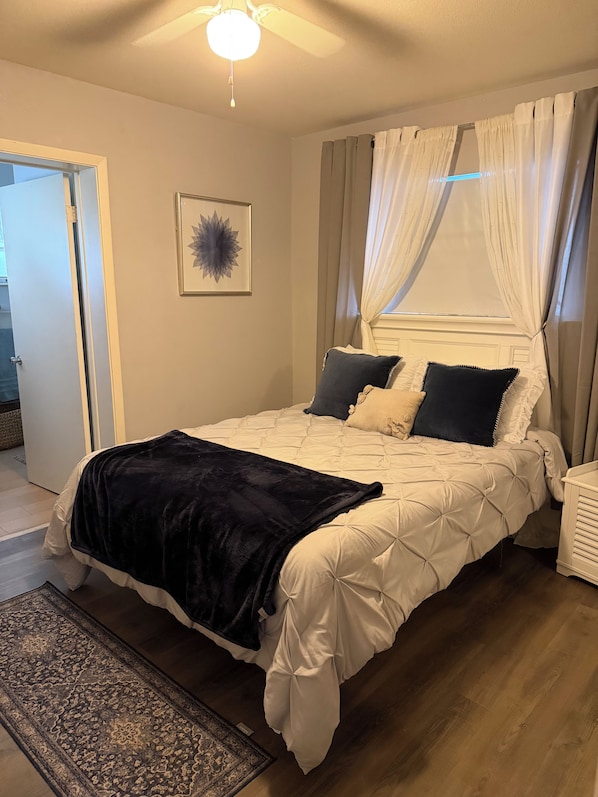 Een slaapkamer met een bed, een plafondventilator, een raam met gordijnen en een vloerkleed.