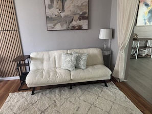 Een sofa in neutrale tinten met patroon kussens, een bijzettafeltje, een vloerlamp en een abstract schilderij in een lijst aan de muur.