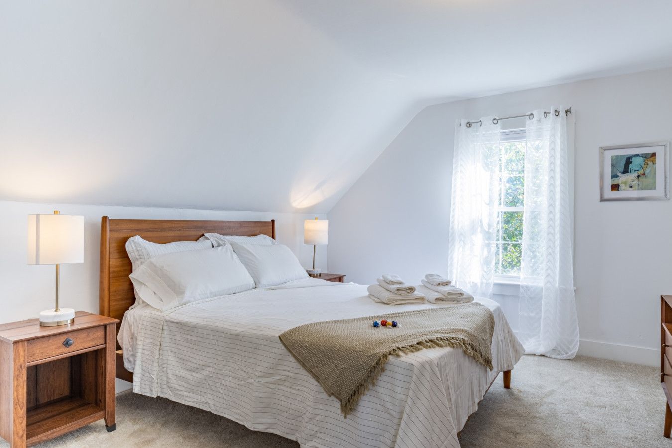 Un dormitorio con cama, mesitas de noche, ventana con cortinas y un cuadro en la pared.