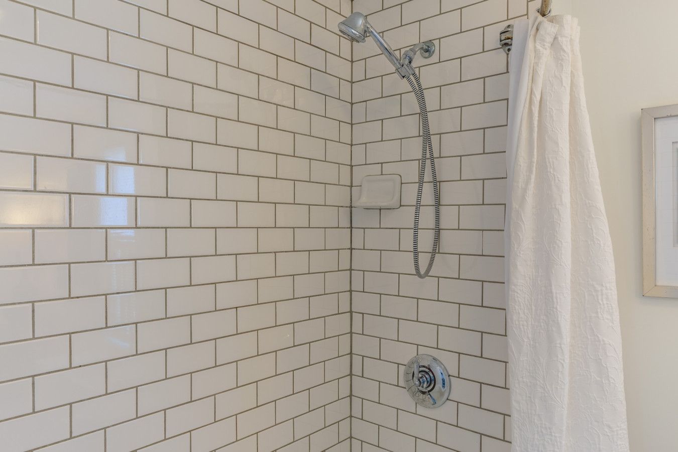 A corner shower with white tiles, a handheld showerhead, and a white shower curtain.