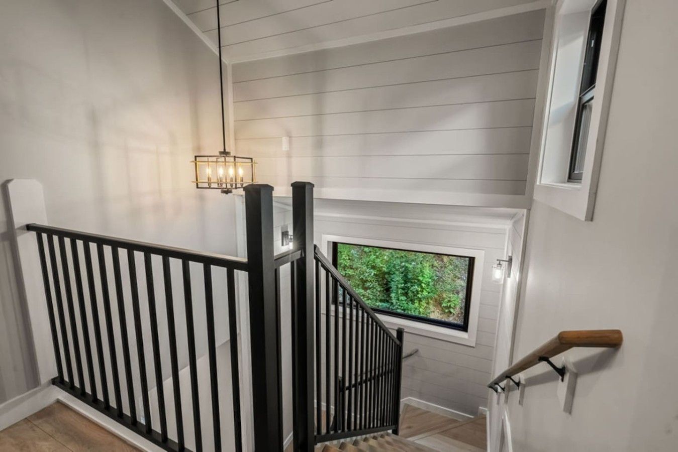 A staircase with a black railing, a hanging light fixture, and a window with a view of greenery.