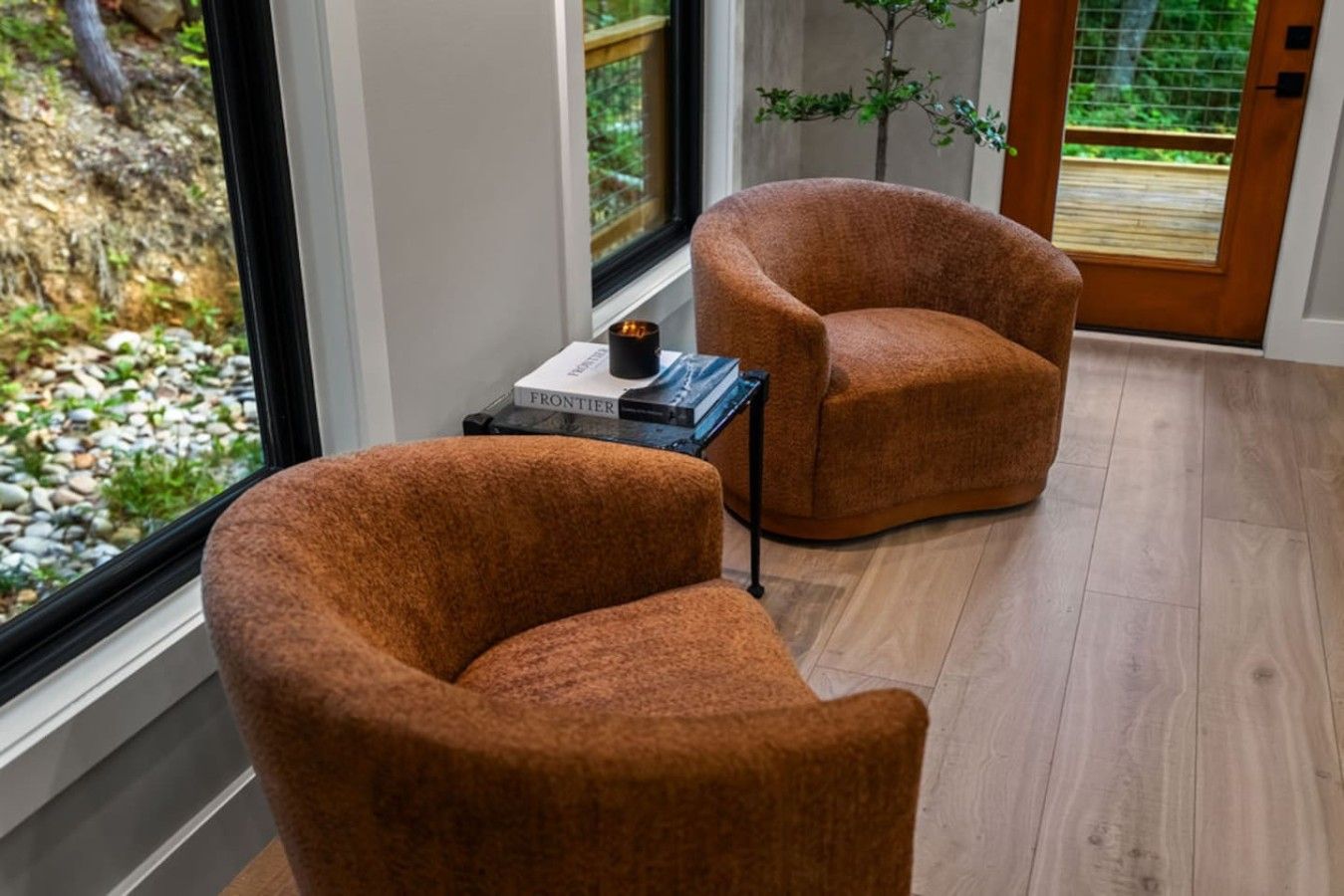 A room with two brown armchairs, a small table with a book and a cup, and a view of a garden through a window.