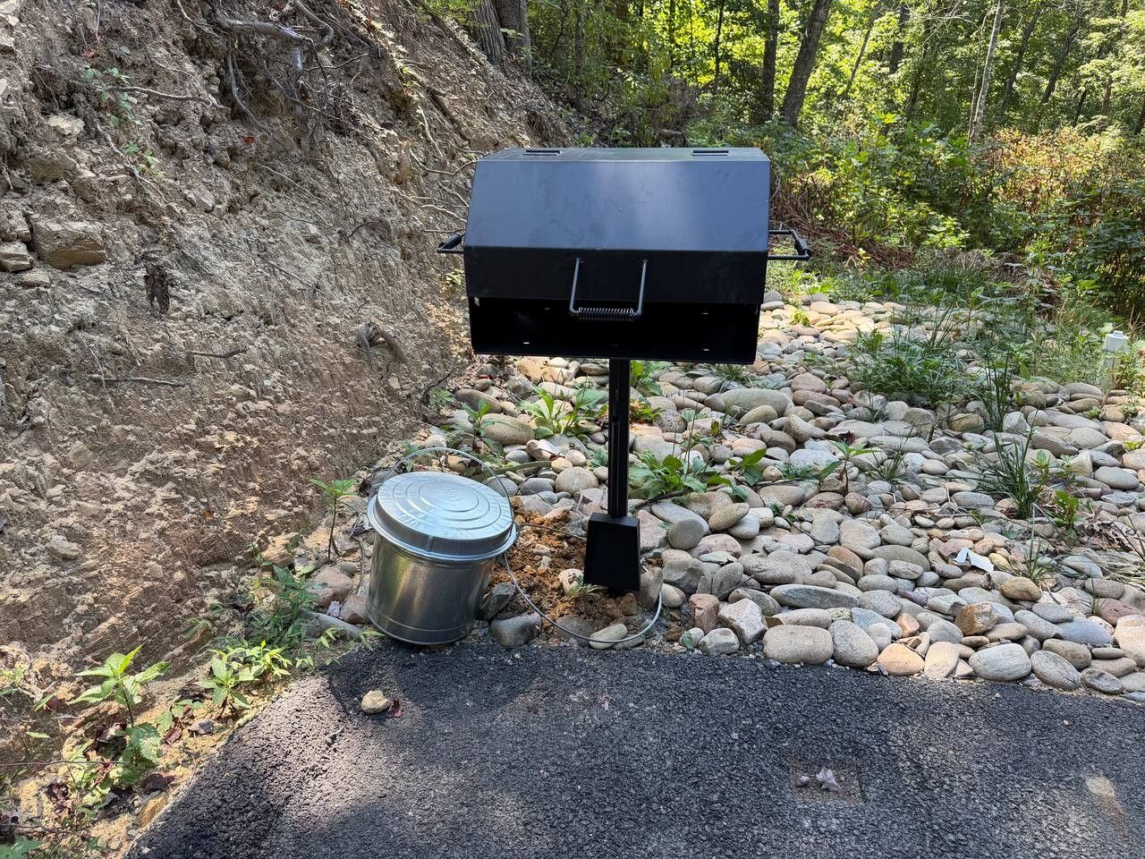 Un barbecue portable, avec un récipient en métal à côté, posé sur un chemin de gravier, avec une pente rocheuse en arrière-plan.