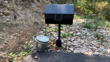 Un barbecue portable, avec un récipient en métal à côté, posé sur un chemin de gravier, avec une pente rocheuse en arrière-plan.