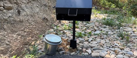 A portable grill with a metal container beside it, set on a gravel path with a rocky slope in the background.