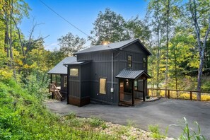 A two-story cabin with a covered porch and a small deck, surrounded by dense forest. - Blackstone Hot Tub • Game Loft (Sevierville)