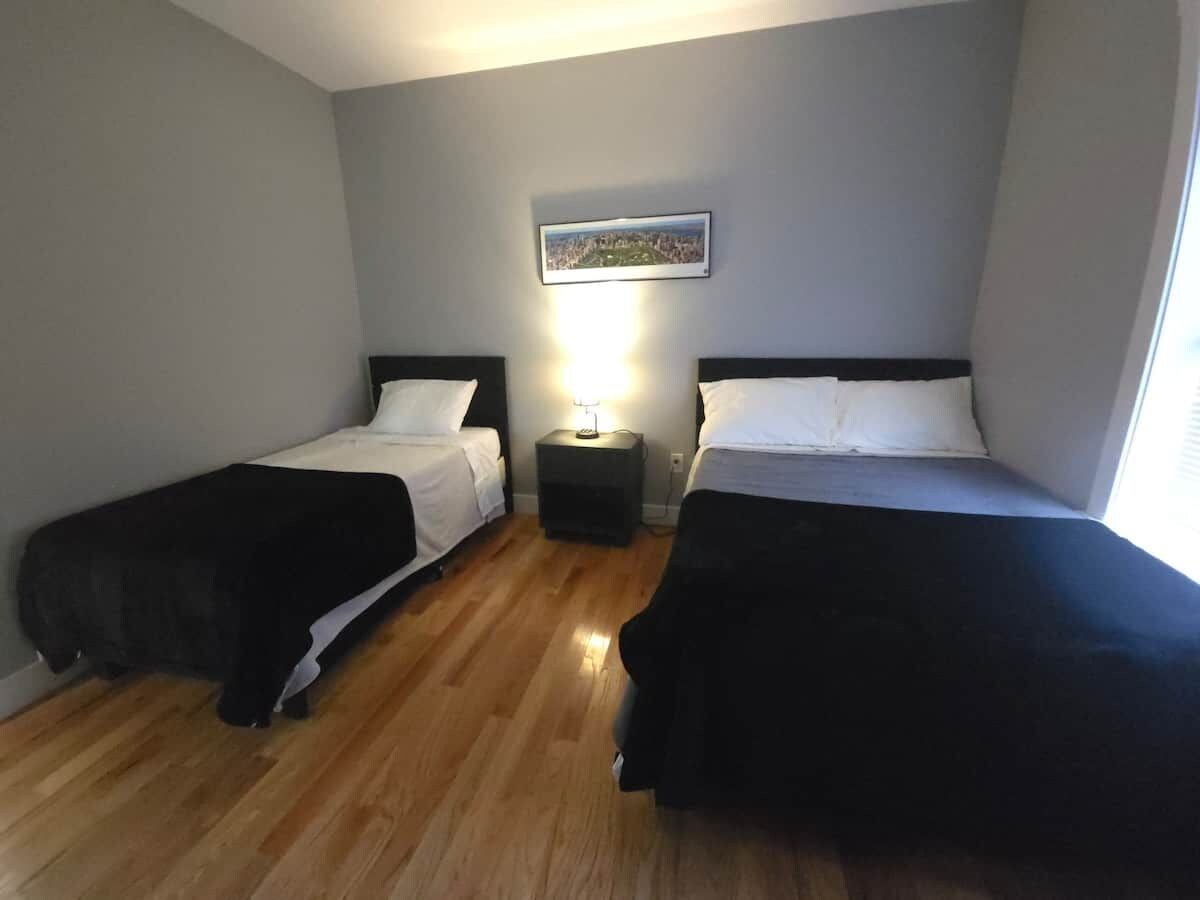 A hotel room with two beds, a nightstand with a lamp, and a cityscape painting on the wall.